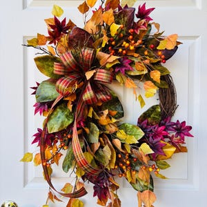May include: A decorative wreath featuring a grapevine base adorned with artificial autumn foliage in shades of yellow, orange, red, and deep green. The arrangement includes small orange berries and a large plaid ribbon bow with long trailing ends.