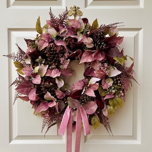 May include: A circular wreath featuring a mix of mauve, burgundy, and muted green foliage, including fern fronds and leaves. The arrangement is accented with dark pinecones and small clusters of berries, finished with two hanging pink velvet ribbons.