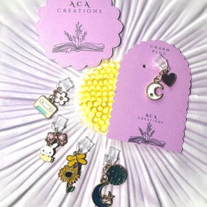 May include: A collection of charm plugs with various designs, including a cassette tape, bunny with a bow, sunflower, and a crescent moon with a star. The plugs are displayed on a white and yellow flower-shaped surface, with two lilac cards that read "ACA CREATIONS".