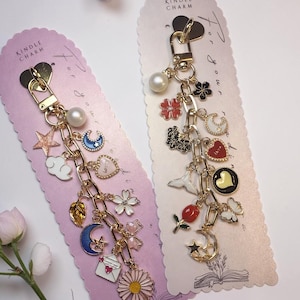 Puede incluir: Dos amuletos Kindle dorados con varios amuletos decorativos, incluyendo lunas, estrellas, flores y corazones. Los amuletos están unidos a una cadena y se muestran en tarjetas rosas con el texto "Kindle Charm".