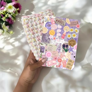 May include: A rectangular notebook with a collage cover featuring purple tones, floral motifs, a smiley face, and text including "MEMORIES" and "TICKET TO HAPPINESS," paired with a thick, hand-crocheted sleeve in pastel pink, yellow, and white yarn.