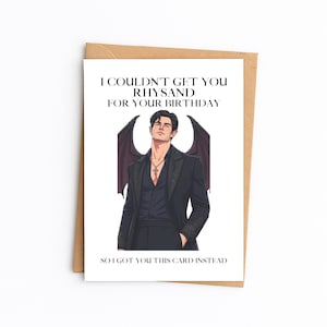 May include: A white greeting card with a brown envelope featuring an illustration of a dark-haired character with wings wearing a black suit and a cross necklace. The text reads: I COULDN'T GET YOU RHYSAND FOR YOUR BIRTHDAY SO I GOT YOU THIS CARD INSTEAD.