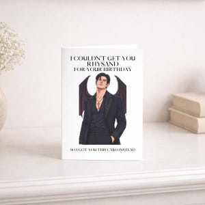May include: A white greeting card featuring a black-haired male character with dark wings, wearing a black suit and necklace. The text reads: I COULDN'T GET YOU RHYSAND FOR YOUR BIRTHDAY SO I GOT YOU THIS CARD INSTEAD. The design is centred.