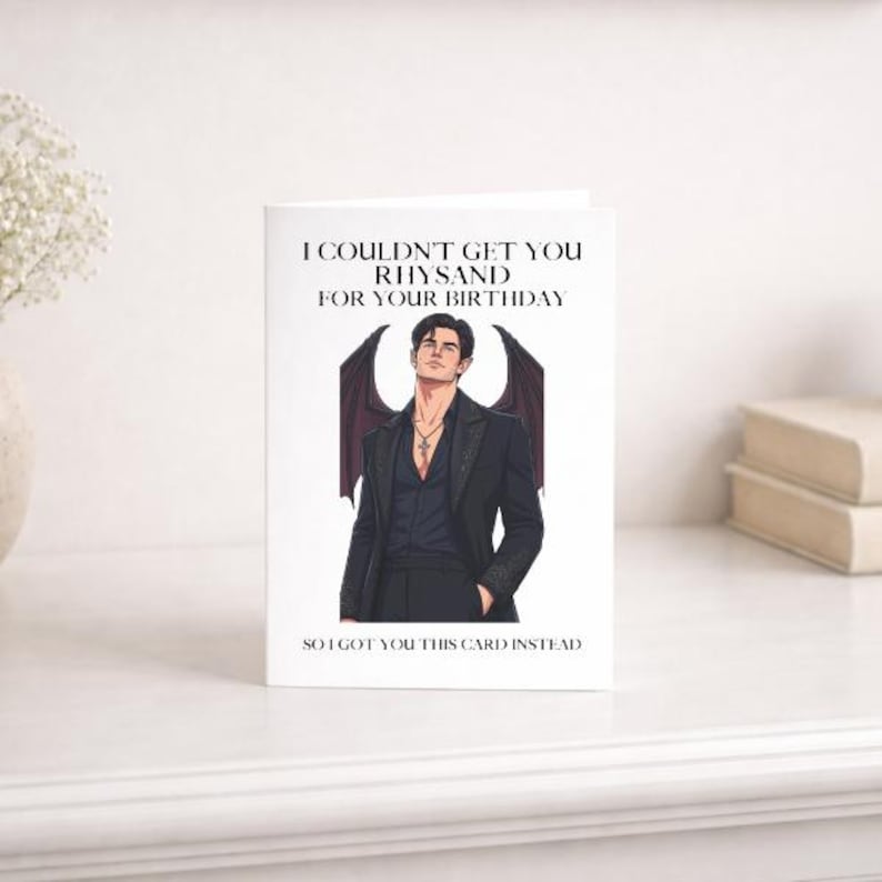 May include: A white greeting card featuring a black-haired male character with dark wings, wearing a black suit and necklace. The text reads: I COULDN'T GET YOU RHYSAND FOR YOUR BIRTHDAY SO I GOT YOU THIS CARD INSTEAD. The design is centred.