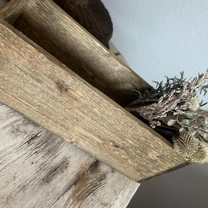 May include: A rustic wooden box with a natural, weathered finish. The box is rectangular with angled sides and contains dried floral arrangements in shades of green, beige, and brown. The box sits on a light-colored wooden surface.