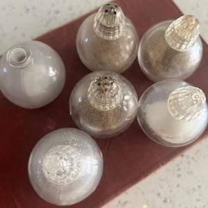 May include: A collection of six clear glass salt and pepper shakers. The shakers are round with clear, textured stoppers. Some shakers contain a light brown substance, while others are empty. The shakers are arranged on a dark red surface.