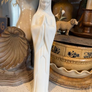 May include: A white ceramic statue of a religious figure with hands clasped in prayer. The figure is wearing a draped robe and a head covering. The statue is standing on a countertop with other decorative items in the background.