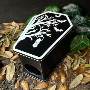 May include: Black coffin-shaped box with a white tree and bat design on the lid. The box has a small opening on the front. The design is reminiscent of Halloween or gothic themes. The box is surrounded by leaves and wood.