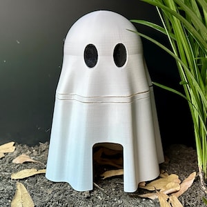 May include: A 3D-printed white ghost figurine with two black oval eyes. The ghost has a draped sheet design with a rectangular opening at the bottom. The figurine is placed on soil with dried leaves, next to green plants.