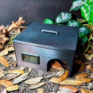 May include: A black reptile enclosure with a textured lid and a small digital thermometer. The enclosure has an arched opening and a handle on top. It is surrounded by dried leaves and greenery, suggesting a natural habitat.