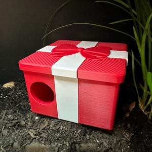 May include: A red pet house, shaped like a gift, with a white ribbon and bow. It features a circular entrance. The textured surface is visible. The house is designed for pets.