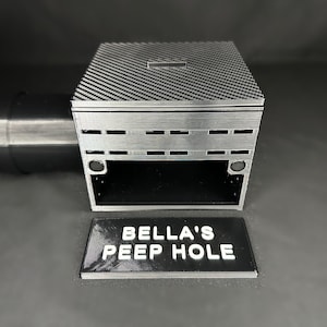 May include: A silver box with a carbon fiber patterned lid and a slot. The front of the box has horizontal rectangular cutouts. A black rectangular sign with the text "BELLA'S PEEP HOLE" is in front of the box.
