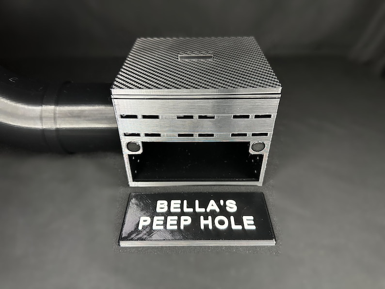 May include: A silver box with a carbon fiber patterned lid and a slot. The front of the box has horizontal rectangular cutouts. A black rectangular sign with the text "BELLA'S PEEP HOLE" is in front of the box.
