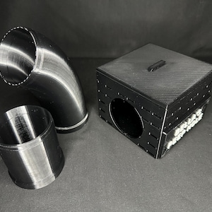 May include: Black 3D-printed components, including a curved pipe, a cylindrical piece, and a box with a circular opening. The box has a small rectangular handle on top and text on the side.
