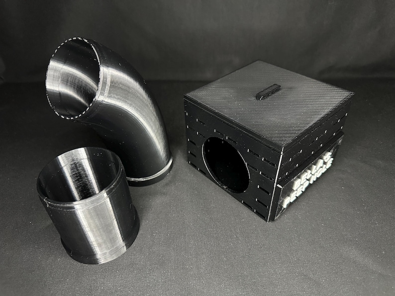 May include: Black 3D-printed components, including a curved pipe, a cylindrical piece, and a box with a circular opening. The box has a small rectangular handle on top and text on the side.