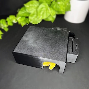 May include: A black, textured, rectangular enclosure with a slot and a small, yellow object visible inside. The enclosure is on a dark surface, with green foliage and a white container in the background.