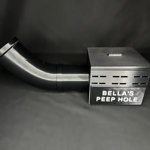 May include: A black, angled tube connected to a rectangular box with a textured top and ventilation slots. The box has a sign that reads "BELLA'S PEEP HOLE". The object is set against a dark background.