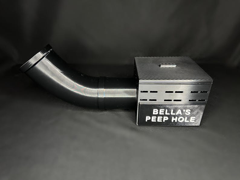 May include: A black, angled tube connected to a rectangular box with a textured top and ventilation slots. The box has a sign that reads "BELLA'S PEEP HOLE". The object is set against a dark background.