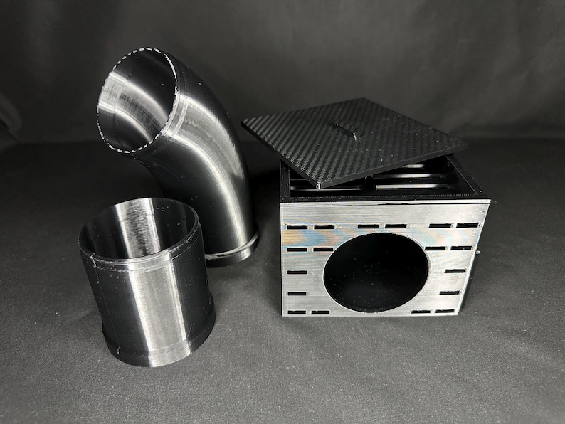 May include: An assortment of metallic components, including a curved pipe, a cylindrical piece, and a square box with a carbon fiber lid. The box has a large circular opening and several rectangular cutouts.