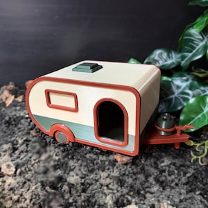 May include: A miniature camper model featuring a cream-coloured body with dark green lower panels and a red trim. The camper includes a small door opening, a rectangular window, a roof vent, and a hitch with a small grey gas cylinder attached.