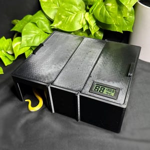 May include: A black rectangular reptile hide box featuring a textured surface and a digital hygrometer display on the top right corner. The device shows a humidity reading of 88 percent. A small yellow snake tail is visible inside the left opening.