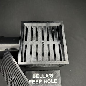 May include: A gray, 3D-printed drain cover with a slotted design, a matching lid, and a sign that reads "BELLA'S PEEP HOLE". A black pipe is attached to the drain.
