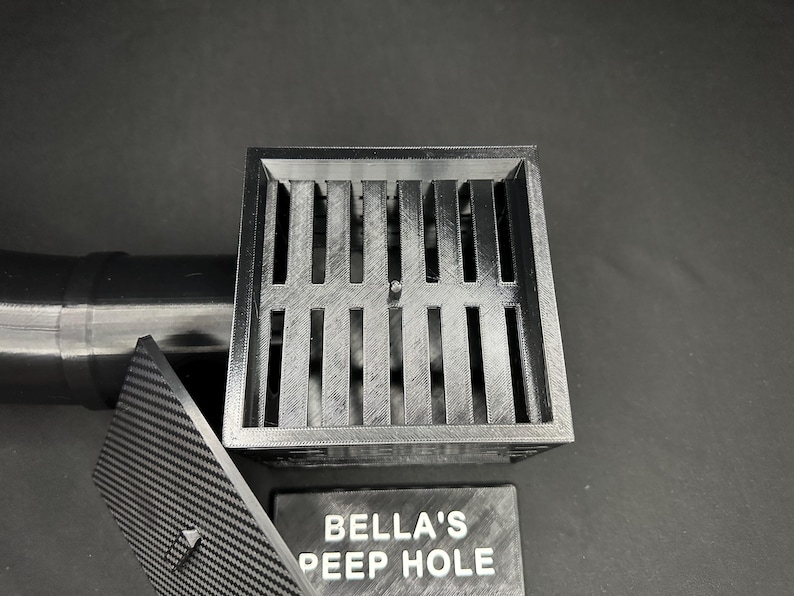 May include: A gray, 3D-printed drain cover with a slotted design, a matching lid, and a sign that reads "BELLA'S PEEP HOLE". A black pipe is attached to the drain.