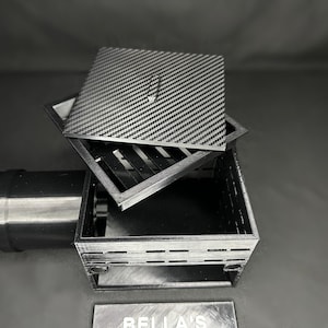 May include: A black and gray square box with a carbon fiber pattern lid. The lid is slightly ajar, revealing the interior of the box. The box is sitting on a dark surface. A metal pipe is visible on the left.