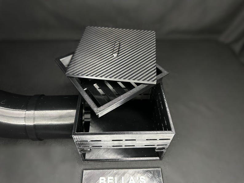May include: A black and gray square box with a carbon fiber pattern lid. The lid is slightly ajar, revealing the interior of the box. The box is sitting on a dark surface. A metal pipe is visible on the left.