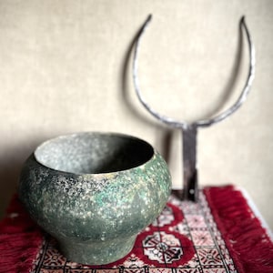 May include: A weathered, spherical green metal bowl with a textured, oxidized patina sits beside a dark, Y-shaped metal tool featuring two upward-curving prongs. The bowl has a wide opening and a tapered base, displaying mottled shades of green.