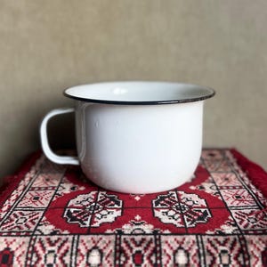 May include: A white enamel mug with a smooth, rounded body and a single side handle. The rim of the mug is finished with a thin, dark-colored metal edge, providing a clean contrast against the glossy white surface of the vessel's exterior.