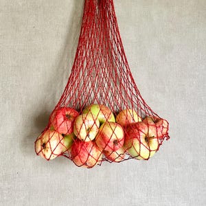 May include: A bright red string mesh market bag hanging vertically, filled with a collection of round apples. The apples display a mix of red and yellow skin tones, visible through the diamond-shaped openings of the knotted cord netting.