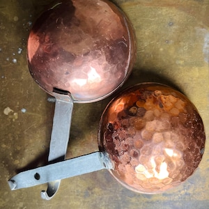 Könnte beinhalten: Zwei runde Kupferschöpflöffel mit gehämmerten Texturen und langen, flachen Griffen aus grauem Metall. Jeder Griff verfügt über ein kleines kreisförmiges Loch am Ende zum Aufhängen. Die Kupferschalen haben eine polierte, reflektierende Oberfläche.