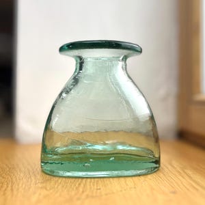Könnte beinhalten: Eine kleine, klare, hellgrüne Glasvase mit einem breiten, abgerundeten Boden, der sich nach oben hin zu einem schmalen Hals und einem flachen, kreisförmigen Rand verjüngt. Das Glas weist dezente, spiralförmige horizontale Linien auf.