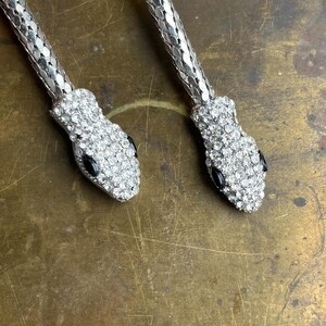 May include: Two silver-toned snake necklaces. The snake heads are encrusted with clear rhinestones and have black eyes. The necklaces are on a brass-colored surface.