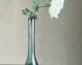 May include: A tall, slender, dark teal glass bud vase with a flared base and a narrow neck. A single white flower with green leaves is positioned inside the vase, with a small white petal resting separately on the surface near the base.