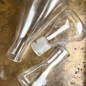 Könnte beinhalten: Drei durchsichtige Laborkolben aus Glas mit konischen Körpern und schmalen Hälsen. Jeder Kolben verfügt über einen Schliffanschluss an der Öffnung. Die Gefäße sind transparent und glatt und zeigen ein klassisches wissenschaftliches Design.