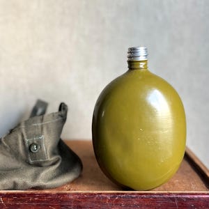 Op de afbeelding: Een olijfgroene veldfles met een glad, rond lichaam en een zilverkleurige metalen schroefdop. Naast de veldfles ligt een gevouwen olijfgroene stoffen tas met een enkele knoopsluiting op een donkerhouten ondergrond.