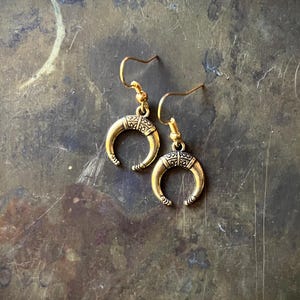 May include: A pair of gold-colored crescent moon earrings. Each earring features a curved, horn-like shape with etched details and a decorative top. The earrings hang from simple gold-toned ear wires, showcasing a bohemian or tribal aesthetic.