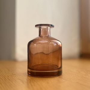 Puede incluir: Una pequeña botella de vidrio translúcido de color ámbar con un cuerpo redondeado y un cuello cilíndrico corto que presenta un borde plano y circular. El vidrio tiene un acabado liso y pulido con un tono marrón cálido y uniforme.