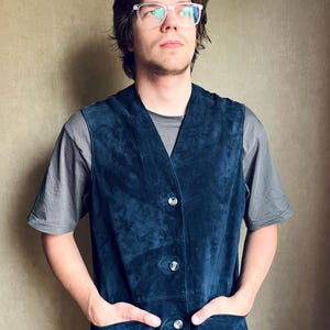 May include: A dark blue suede vest featuring a V-neckline, four front buttons, and two side pockets, worn over a plain gray short-sleeved t-shirt. The vest has a soft, matte texture and a structured fit with visible stitching along the edges.