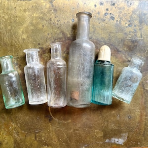 Può includere: Una collezione di sei bottiglie di vetro di varie forme e dimensioni. Quattro bottiglie sono in vetro trasparente, una è una bottiglia alta e leggermente torbida, e una è una bottiglia rettangolare in vetro verde acqua con un tappo bianco arrotondato.