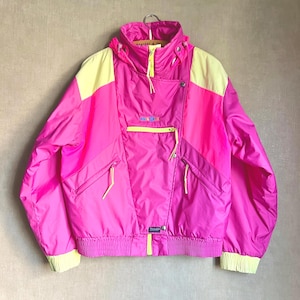 May include: A bright pink and yellow windbreaker jacket featuring a high collar, a hood, and multiple zippered pockets on the front. The jacket includes a central chest pocket with a small rectangular logo patch and ribbed cuffs at the sleeves.