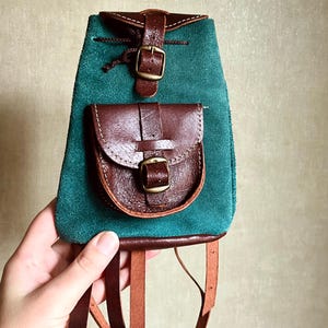 Pode incluir: Uma mochila pequena de camurça verde-azulada com detalhes em couro castanho escuro, incluindo um bolso frontal com fecho de fivela, uma aba superior com correia de fivela e alças finas de couro castanho presas à base da mala.