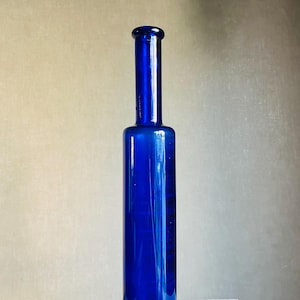Op de afbeelding: Een hoge, cilindrische glazen fles in een levendige kobaltblauwe kleur. De fles heeft een glad, recht lichaam dat overgaat in een slanke, langwerpige hals, bekroond met een eenvoudige, afgeronde opening aan de bovenzijde.