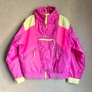 May include: A bright pink and yellow color-blocked jacket featuring a high collar, a hood, and long sleeves with ribbed cuffs. The jacket includes a central zippered pocket, two side zippered pockets, and a front zipper closure with yellow pulls.