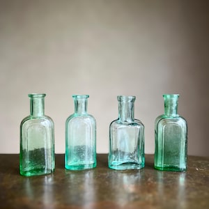 Könnte beinhalten: Vier kleine, durchscheinende hellgrüne Glasflaschen, die in einer Reihe angeordnet sind. Jede Flasche hat einen rechteckigen Körper mit abgerundeten Schultern, einen kurzen Hals und eine schmale Öffnung mit Rand. Das Glas wirkt leicht texturiert und antik.