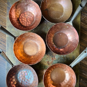 May include: A collection of six round copper pans with hammered textures and metal handles. Each pan features a distinct reddish-brown metallic finish, with some showing signs of oxidation and patina. The pans are arranged in a cluster.