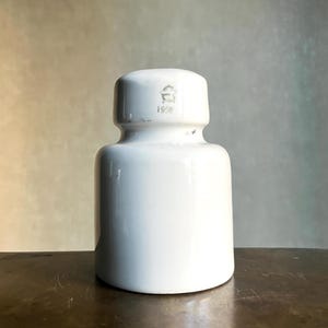 Pode incluir: Um isolador elétrico de cerâmica branca com corpo cilíndrico e seção superior escalonada. A parte superior apresenta um logótipo estampado e o ano 1958. O objeto tem um acabamento liso e brilhante com um pescoço distinto que separa a base.