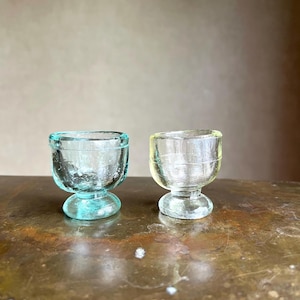 May include: Two small glass egg cups with a rounded bowl and a thick, circular pedestal base. One cup is a translucent light blue-green, while the other is a clear, pale yellow. Both items feature a textured, slightly uneven surface finish.